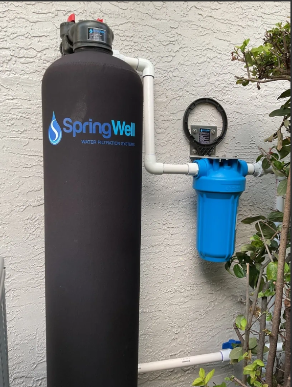SpringWell whole house water filtration system installed on exterior wall featuring large black main tank with blue logo and connected blue pre-filter housing, designed for comprehensive home water purification"RetryClaude can make mistakes. Please double-check responses.