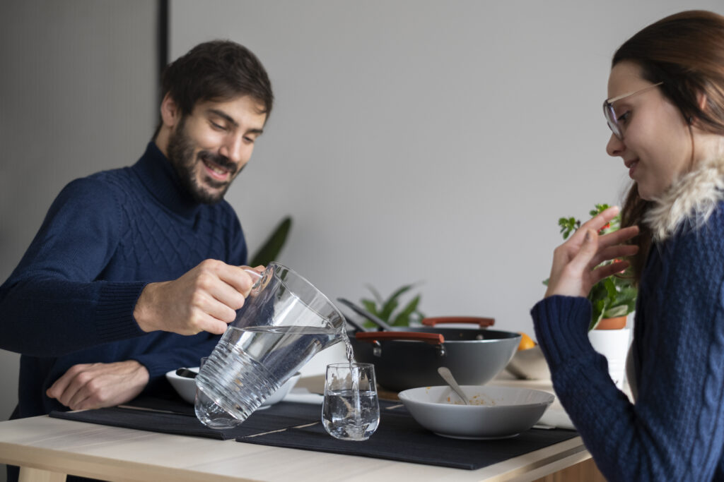 Couple enjoying a meal at home with man pouring clear filtered water from a glass pitcher into drinking glasses, highlighting the benefits of home water filtration systems